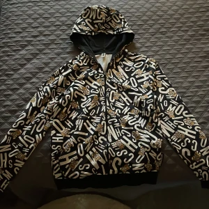 Moschino hoodie med björnar och tryck - Svart hoodie från Moschino med vit textprint och bruna björnar över hela jackan. Jackan har huva, dragkedja framtill och långa ärmar. Perfekt för dig som gillar streetwear och coola detaljer.