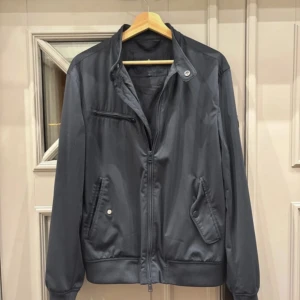 Marinblå jacka från Morris Stockholm S -  Jacka från Morris Stockholm i storlek S. Jackan har dragkedja framtill, ståkrage med knapp, tre fickor med detaljerade knappar och en diskret logotyp på ärmen. Perfekt för dig som gillar stilren och sportig look.