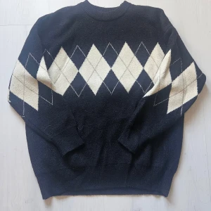 Svart stickad tröja med argylemönster - Svart stickad tröja med klassiskt argylemönster i vitt och beige över bröstet och ärmarna. Tröjan har rund halsringning och långa ärmar, perfekt för dig som gillar preppy vibes och vill sticka ut lite extra.