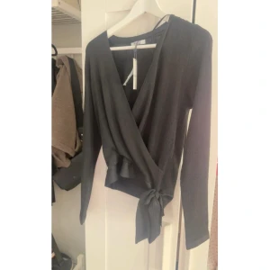 Svart omlottopp med knyt i midjan - Snygg svart omlottopp med långa ärmar och knyt i midjan. Toppen har en djup v-ringning och är tillverkad i ett mjukt, ribbat material som ger en skön känsla. Perfekt för dig som gillar stilrena och trendiga plagg.