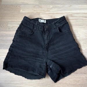 Svarta jeansshorts från Gina Tricot - Svarta jeansshorts från Gina Tricot med råa kanter och klassisk femficksdesign. Hög midja och knappstängning framtill. Snygg, enkel look som passar perfekt till sommaren. 