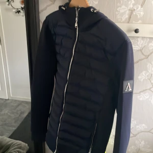 Mörkblå cardiganjacka Alessandro Zavetti - Snygg mörkblå cardiganjacka från Alessandro Zavetti med huva och dragkedja framtill. Jackan har quiltad front, släta ärmar och en patch med logga på vänster arm. Perfekt passform och stilren look för dig som vill ha något fräscht och modernt.