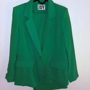 Grön kostym från JDY, storlek M - Snygg grön kostym från JDY i storlek M. Kavajen har klassisk krage, en knapp framtill och två fickor med lock. Byxorna är raka och matchar kavajen perfekt. Materialet är mjukt och lätt polyester, vilket gör den bekväm att bära. Perfekt för dig som vill sticka ut med färg.