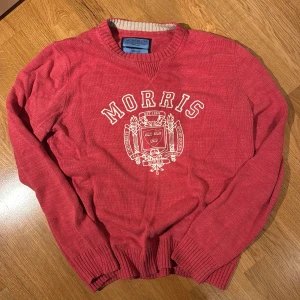 Röd stickad tröja från Morris Co - Säljer en röd stickad tröja från Morris Co med vit broderad logga och emblem på bröstet. Tröjan har rund halsringning, ribbade muddar och lång ärm. Perfekt för dig som gillar klassisk college-stil.