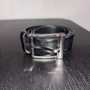 Svart skärp i skinn med silverfärgat spänne - Klassiskt svart bälte i skinn med ett stilrent, fyrkantigt silverfärgat spänne. Perfekt accessoar för att lyfta din outfit med en enkel och tidlös look. Bältet har en slät yta och passar till de flesta byxor.