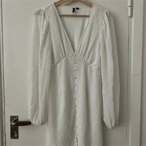 Vit långklänning med knappar och v-ringning - Superfin vit klänning med lång ärm och v-ringning. Klänningen har dekorativa knappar framtill och ett diskret prickigt strukturmönster i tyget. Dubbel tyg vilket gör att den inte ska vara see thru. Lätt puffade axlar och resår vid ärmslut ger en romantisk vibe. Har använts en gång och har en pytte liten fläck på övre delen av ryggen. Är dock perfekt för tex vår och sommar!🤍