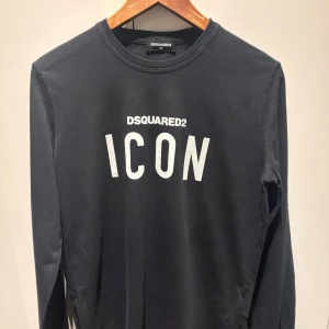 Svart långärmad tröja DSQUARED2 ICON - Svart långärmad tröja från DSQUARED2 med vit 'ICON'-text på bröstet. Klassisk rund halsringning och stilren design, perfekt för dig som gillar streetwear och vill sticka ut med en ikonisk look. Tillverkad i mjuk bomull för skön känsla hela dagen.