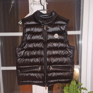 Svart dunväst från Moncler - Snygg svart dunväst från Moncler med glansig finish och klassisk logga på bröstet. Västen har ståkrage, två fickor med dragkedja och quiltad design. Perfekt för lager-på-lager och har en modern, stilren look. Dragkedja framtill och insida med märkning.