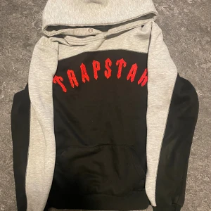 Trapstar hoodie i svart och grå - Säljer en snygg hoodie från Trapstar i svart och ljusgrått med röd broderad logga på bröstet. Tröjan har huva, magficka och långa ärmar. Perfekt för dig som gillar streetwear och vill sticka ut med cool design.