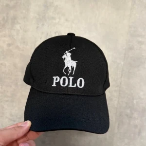 Svart keps från Polo Ralph Lauren - Snygg svart keps från Polo Ralph Lauren med klassisk broderad logga i vitt framtill. Justerbar rem baktill med metallspänne för perfekt passform. Kepsen är tillverkad i bomull och har en böjd skärm.