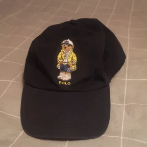 Keps Polo Ralph Lauren björn - Svart keps från Polo Ralph Lauren med broderad björn i gul jacka framtill och gul POLO-text. Justerbar rem med metallspänne bak och broderad logga. Tillverkad i 100% bomull. Klassisk böjd skärm och avslappnad passform.