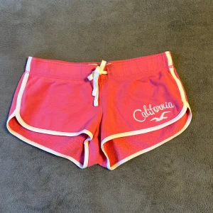 Rosa Hollister shorts med snörning XS - Snygga rosa shorts från Hollister med vit kant och broderad 'California'-text framtill. De har snörning i midjan och är gjorda i mjuk bomull, perfekta för chill dagar. Sportig och loose passform med klassisk Hollister-vibe.