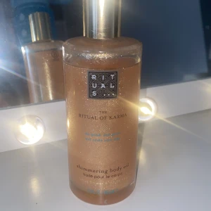Rituals The Ritual of Karma Shimmering Body Oil - Shimmering body oil från Rituals i serien The Ritual of Karma. Oljan har en guldig, skimrande finish och innehåller lotusblomma och vitt te. Kommer i en genomskinlig flaska med guldfärgat lock och ger huden en snygg glow. 