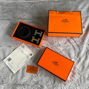 Svart H bälte med H-spänne - Exklusivt svart bälte från H med två utbytbara H-spännen i silver och guld. Bältet är stilrent och tillverkat i äkta skinn, levereras i original orange box med kvitto och certifikat. Perfekt accessoar för dig som vill sticka ut med lyxig känsla.