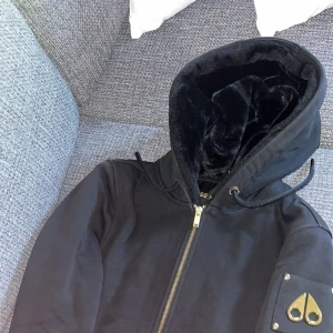  Moose Knuckles Bunny hoodie - Svart pufferjacka från Moose Knuckles med mjukt pälsfoder i huvan, guldfärgad dragkedja och ikonisk logga i metall på ärmen. Jackan har snörning i huvan och praktiska fickor framtill. Perfekt för kalla dagar och riktigt snygg streetstil.