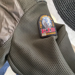 Svart dunväst från Parajumpers - Säljer en svart dunväst från Parajumpers med huva, quiltad framsida och coolt PJS-märke på axeln. Västen har dragkedja framtill, bröstficka med dragkedja och en gul detalj vid halsen. Perfekt för lager-på-lager och streetstyle.