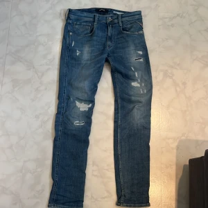 Replay Anbass blå jeans slim fit - Snygga Replay Anbass jeans i blå tvätt med slitningar och detaljerade sömmar. Slim fit-modell med klassisk femficksdesign, knapp och dragkedja i midjan samt läderpatch bak. Perfekt för dig som gillar en modern och avslappnad look.