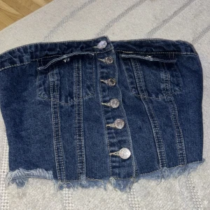 Mörkblå denim tubtopp från SHEIN - Säljer en mörkblå denim tubtopp från SHEIN med snygga silverfärgade knappar framtill, två fejkade bröstfickor och rå, fransig kant nertill. Toppen har smockad rygg för extra passform och är tillverkad i bomullsmix. Aldrig använd