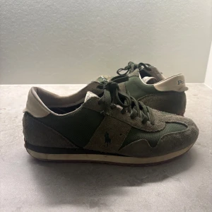Gröna sneakers från Polo Ralph Lauren - Snygga sneakers från Polo Ralph Lauren i mörkgrönt och beige. Ovandel i mocka och textil med klassisk Polo-logga på sidan och hälen. Skorna har snörning och en brun gummisula med grepp. Perfekta för en avslappnad och sportig look.