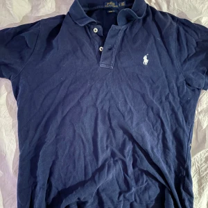 Marinblå pikétröja från Ralph Lauren - Klassisk marinblå pikétröja från Polo Ralph Lauren i skinny fit. Tröjan har korta ärmar, krage och knäppning med tre knappar framtill. Ikoniska vita loggan broderad på bröstet. Perfekt för en stilren och sportig look.