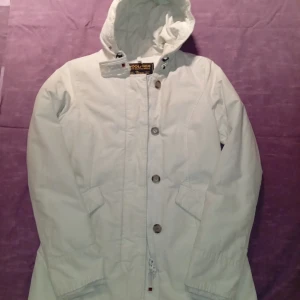 Vit dunjacka med huva från Woolrich - Snygg vit dunjacka från Woolrich med huva och stora knappar framtill. Jackan har två snedställda fickor och är fodrad för extra värme. Perfekt för kalla dagar och har en klassisk, stilren look. 60/40 Arctic Parka Slim. Ovanligare färgen vit.