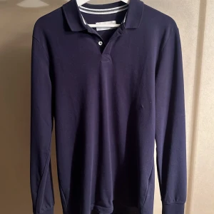 Mörkblå långärmad pikétröja - Säljer en stilren mörkblå långärmad pikétröja med klassisk krage och knäppning framtill. Tröjan är i mjuk bomull och passar perfekt till både jeans och chinos. Enkel och tidlös design som funkar året runt.