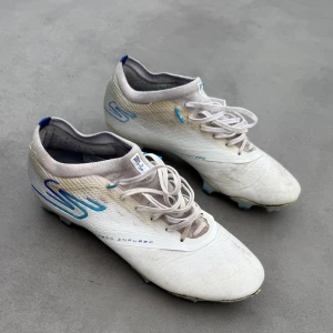 Sketchers Diamond Ice - vita och blåa fotbolsskor - Säljer ett par vita Diamond Ice Sketchers   fotbollsskor med blå detaljer och snörning. Skorna har en smidig och lätt design med en rund tå, perfekt för snabba rörelser på planen.