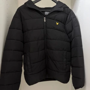 Svart pufferjacka från Lyle & Scott - Svart pufferjacka från Lyle & Scott med huva och gul logotyp på bröstet. Jackan har quiltad design, dragkedja framtill och två sidofickor. Perfekt för kalla dagar och har en clean, stilren look som funkar till det mesta. Pris kan diskuteras.