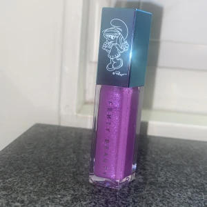 Fenty Beauty Gloss Bomb Smurfette - Fenty Beauty Gloss Bomb i färgen Smurfette Effect, ett glittrigt lila läppglans med coolt Smurfan-motiv på locket. Kommer i en snygg fyrkantig förpackning med metallicblått lock och transparent hylsa som visar den skimrande lila nyansen. Perfekt för dig som gillar unika limited edition-produkter.