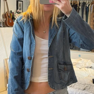 Jeansjacka med knappar och fickor - Säljer en snygg jeansjacka i klassisk blå denim med två stora fickor framtill och bruna knappar. Jackan har en croppad passform och markerad krage, perfekt att slänga över en t-shirt. Enkel och stilren design som funkar till det mesta.