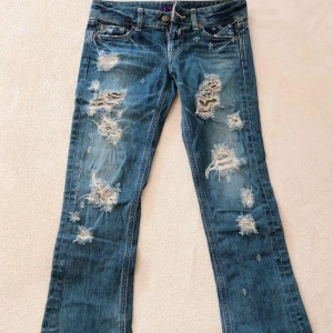 Japanska lågmidjade Jeans  - Riktigt snygga blå jeans med bootcut passform och rejält slitna detaljer framtill. Jeansen har klassisk femficksdesign, dragkedja och coola lila etiketter. Perfekta för dig som gillar en edgy och avslappnad stil. Midja-35cm och grenmått-18 cm