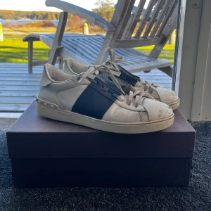 Valentino Garavani Open sneakers vit/blå - Säljer ett par Valentino Garavani Open sneakers i vitt skinn med bred blå läderdetalj över mitten. Skick 8/10, inga skador bara använd. Storlek 44, kvitto finns, allt og på bilderna följer med :). Pris går att diskutera✅