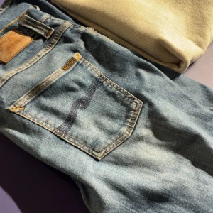 Blå Nudie Jeans - Säljer ett par klassiska blå jeans från Nudie Jeans med snygga slitningar (33/32). Jeansen har straight/slim passform och svart broderad bakficka. Jeansen har några få mindre defekter; hål i vänstra framfickan, hål på ena knät och ett litet hål/slitning i skrevet. Inga större problem dock då det är helt gratis att laga hos Nudie Jeans. Hör gärna av er vid frågor!