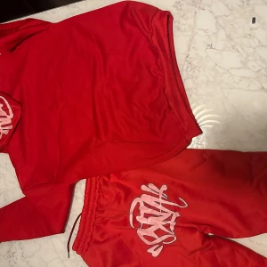 Röda mjukisbyxor med tryck - Röda mjukisbyxor med resår och snörning i midjan samt stort vitt tryck på ena benet. Byxorna har en avslappnad passform och är tillverkade i mjukt material, perfekta för chill dagar eller träning.