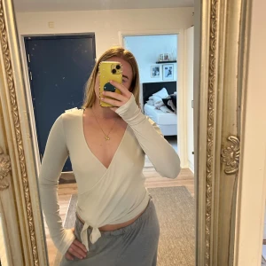 Pilateströja - Säljer en beige/vit omlottopp från Even&Odd Active med lång ärm och djup v-ringning. Toppen har knyt i midjan och är tillverkad i ett mjukt, stretchigt material som sitter snyggt på kroppen. Perfekt för dig som gillar stilrena och trendiga plagg. Säljer pga för stor!