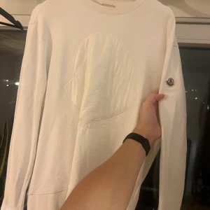 Vit sweatshirt från Moncler - Säljer en stilren vit sweatshirt från Moncler med stort ton-i-ton logomärke på bröstet och klassisk patch på ärmen. Tröjan har rund hals och långa ärmar, perfekt för dig som gillar exklusiva och cleana plagg.