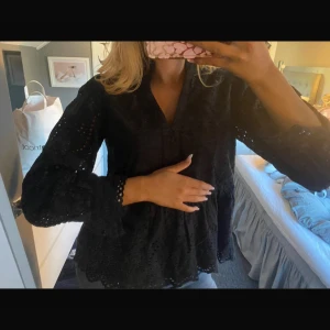 Svart blus med spetsdetaljer - Säljer en svart blus med v-ringning och långa ärmar. Blusen har fina spetsdetaljer och broderade hålmönster som ger en cool och feminin vibe. Ärmarna är lätt puffade och avslutas med spetskant. Perfekt för dig som gillar stilrena och detaljerade toppar.