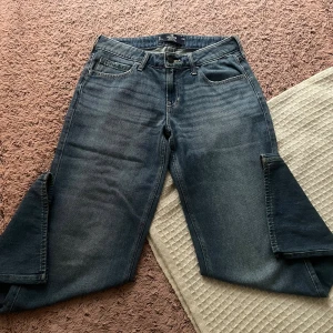 Mörkblå jeans från Hollister - Snygga mörkblå jeans från Hollister med klassisk femficksdesign och brunt läderpatch bak. Jeansen har rak passform och subtila slitningar framtill för en cool look. Passformen är även baggy om man har storlek Xs-M.