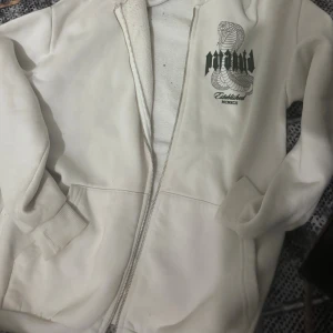 Vit hoodie med tryck från Paranoia - Vit hoodie med dragkedja och huva från Paranoia. På bröstet finns ett stort tryck med en kobramotiv och texten 'Paranoia Established MCMXCII'. Mjuk insida och ribbade muddar vid ärmslut och nederkant. Perfekt för dig som gillar streetwear och coola prints.