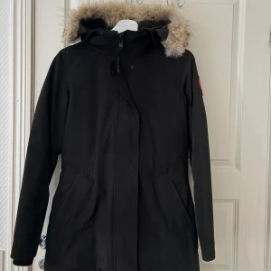 Svart parkas från Canada Goose med päls - Säljer en svart Victoria parkas från Canada Goose med klassisk patch på ärmen och fluffig päls runt huvan. Jackan har dragkedja framtill, två stora fickor och är perfekt för kalla vinterdagar. Materialet är slitstarkt och isolerande, designad för att hålla dig varm. Kan diskutera pris vid snabbaffär!