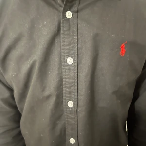 Svart skjorta från Ralph Lauren - Klassisk svart skjorta från Ralph Lauren med röd broderad logga på bröstet. Skjortan har knappar framtill och en stilren krage. Tillverkad i bomull för en skön känsla och snygg passform.