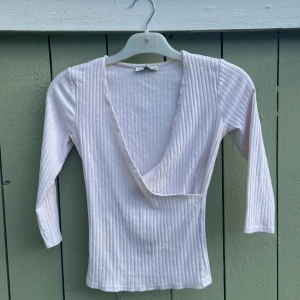 Vit ribbad omlottopp från Bershka - Säljer en vit ribbad omlottopp från Bershka i storlek M. Toppen har djup v-ringning, trekvartsärm och är tillverkad i mjukt bomullsmaterial. Perfekt för dig som gillar stilrena och enkla plagg med en twist.