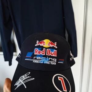 Red Bull Racing keps Pepe Jeans - Snygg svart Red Bull Racing keps från Pepe Jeans London med broderade loggor, nummer 1 och autograf på skärmen. Kepsen har justerbar rem baktill och detaljer i vitt, blått och rött. Perfekt för dig som älskar Formel 1 och motorsport.