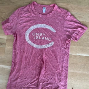 Rosa Coney Island t-shirt från Alternative - Snygg rosa t-shirt från Alternative med stort vitt Coney Island-tryck på bröstet. T-shirten har rund hals och korta ärmar, tillverkad i mjuk bomullsblandning som känns skön mot huden. Perfekt för dig som gillar streetwear och grafiska prints.