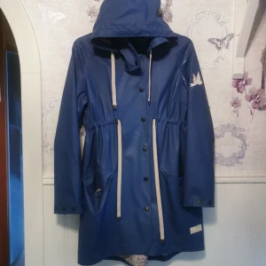 Blå regnjacka från Odd Molly med huva - Blå regnjacka från Odd Molly med huva och dragsko i midjan. Jackan har stora fickor framtill, tryckknappar och ett snyggt Odd Molly-tryck på ärmen. Insidan har ett mönstrat foder som ger extra stilpoäng. Perfekt för regniga dagar! Den har några en stacka fläckar på sig, har ej testat ta bort.. 