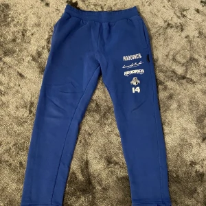  Hoodrich mjukisbyxor - Snygga blå mjukisbyxor från Hoodrich med flera vita tryck och broderade detaljer på ena benet, bland annat logga och siffran 14. Byxorna har snörning i midjan och fickor på sidorna. Perfekt för chill eller streetwear.