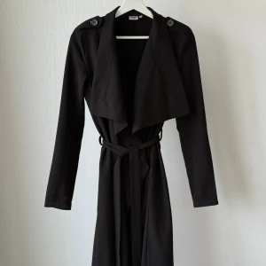 Fin svart trenchcoat från Object i XS - Svart trenchcoat från Object i 100% polyester. Jackan har långa ärmar, bred krage, axelklaffar med knappar och knytskärp i midjan. Snygg och stilren modell med klassiska detaljer och rak passform.