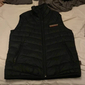 Svart dunväst från Napapijri - Svart quiltad dunväst från Napapijri med dragkedja framtill och två fickor med dragkedja. Västen har hög krage och en broderad logotyp på bröstet. Tillverkad av minst 70% återvunnet material, perfekt för lager-på-lager-stil. Helt oanvänd 