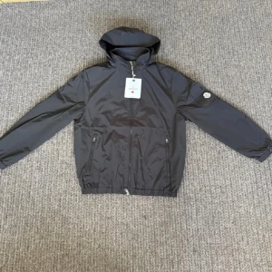 Svart vindjacka från Moncler med huva - Svart vindjacka från Moncler med huva, hel dragkedja och två fickor med dragkedja framtill. Jackan har elastisk midja och ärmar, samt en ficka med Moncler-logga på ärmen. Tillverkad i lätt och slitstark polyester, perfekt för blåsiga dagar.