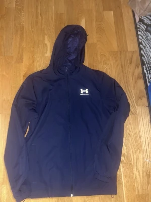 Marinblå vindjacka från Under Armour - Säljer en marinblå vindjacka från Under Armour med huva och hel dragkedja framtill. Jackan har en enkel och sportig design med logga på bröstet och elastiska muddar vid ärmsluten. Perfekt för blåsiga dagar eller till träningen.